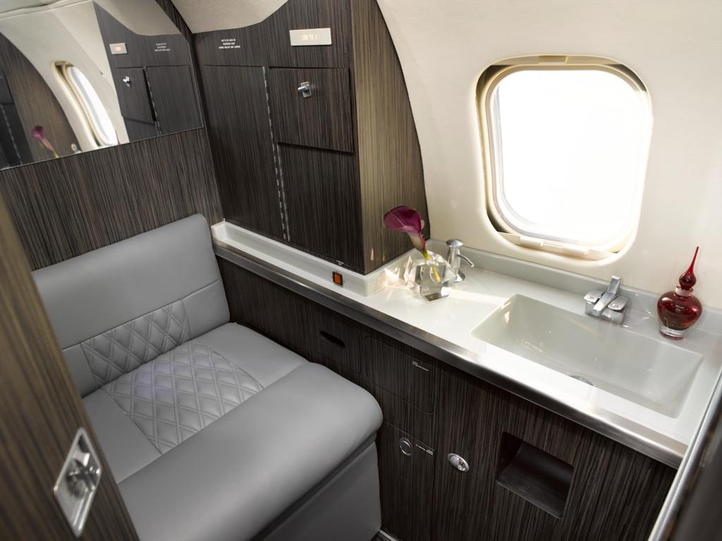 Aircraft lavatory with an upholstered seat, white vanity, integrated basin, mirror, and dark cabinetry.